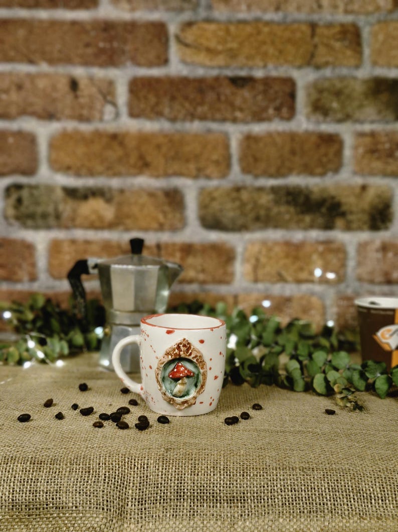 May include: A white ceramic mug with a red rim and a mushroom design. The mug has a handle and is decorated with a framed mushroom illustration. Coffee beans are scattered around the mug, with a brick wall in the background.