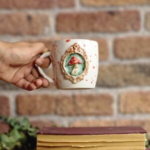 May include: White ceramic mug with a whimsical mushroom design. The mug features a raised, framed mushroom motif in red, green, and brown. Red speckles dot the mug's surface. The mug is set against a brick background and rests above two books.