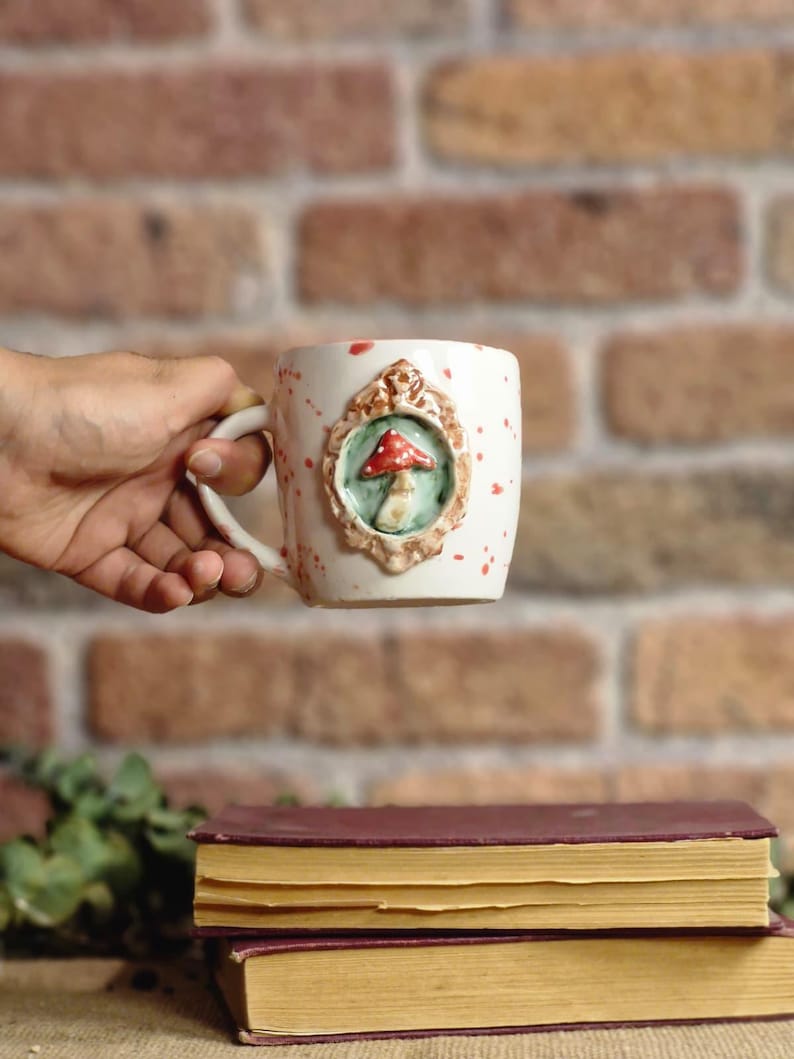 May include: White ceramic mug with a whimsical mushroom design. The mug features a raised, framed mushroom motif in red, green, and brown. Red speckles dot the mug's surface. The mug is set against a brick background and rests above two books.