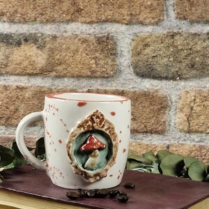 May include: A white ceramic mug with a mushroom design. The mug has a handle and is decorated with red speckles. A raised, framed mushroom is centered on the mug. The mug is sitting on a stack of books.