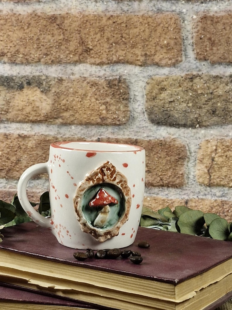 May include: A white ceramic mug with a mushroom design. The mug has a handle and is decorated with red speckles. A raised, framed mushroom is centered on the mug. The mug is sitting on a stack of books.