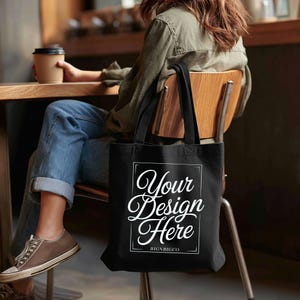 May include: A black canvas tote bag featuring a white rectangular border and the white text "Your Design Here" with "BIGNBIGCO" printed underneath in a smaller font. The bag has two black shoulder straps and a flat rectangular base.