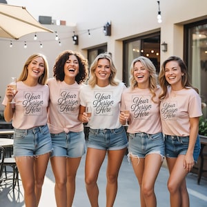 May include: Five women wearing light pink and white t-shirts with the text "Your Design Here" in a black frame. They are wearing denim shorts and holding glasses of champagne. The scene is outdoors with string lights.