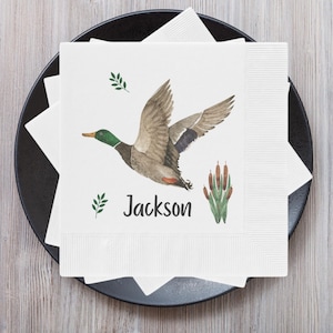 May include: White paper napkin with a watercolor illustration of a green-headed mallard duck flying over green cattails. The napkin has the name "Jackson" printed in black below the duck.