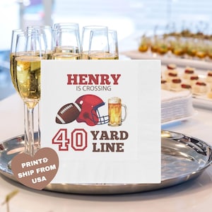 May include: White cocktail napkins with a football theme. The napkins feature a football, a red helmet, and a mug of beer. Text on the napkin reads "HENRY IS CROSSING 40 YARD LINE". Champagne flutes and appetizers are in the background.