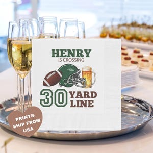 Könnte beinhalten: Weiße quadratische Serviette mit Football-Motiv. Die Serviette zeigt einen Football, einen Helm und ein Bierkrug-Motiv sowie den Text "HENRY IS CROSSING 30 YARD LINE". Die Serviette liegt auf einem silbernen Tablett.