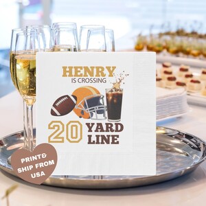 May include: White square napkin with a football, helmet, and drink graphic. Text reads "HENRY IS CROSSING 20 YARD LINE." The napkin is on a silver tray with champagne flutes in the background. A heart-shaped sticker says "PRINT & SHIP FROM USA."