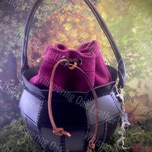 May include: A black leather bucket bag with a dark magenta fabric drawstring insert. The bag features a single arched handle, stitched panel construction, and a silver-toned metal charm with a crescent moon and beads attached to the side.