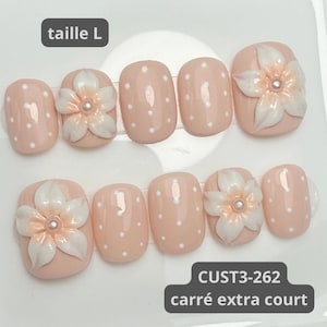 Könnte beinhalten: Ein Set aus zehn künstlichen Fingernägeln in hellem Pfirsichton mit weißen Punkten. Einige Nägel sind mit einer dreidimensionalen weißen Blume mit einer kleinen Perle in der Mitte verziert. Das Bild enthält die Texte taille L, CUST3-262 und carré extra court.