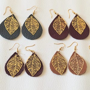 Peut inclure: Huit paires de boucles d'oreilles en forme de goutte disposées sur deux rangées. Chaque boucle présente une base texturée de différentes couleurs, dont noir, rouge foncé, marron et gris, surmontée d'une breloque feuille dorée et d'un crochet.