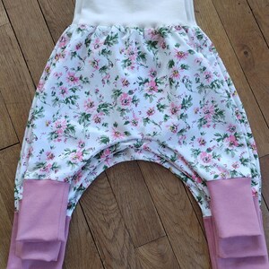 Peut inclure: Pantalon de bébé à imprimé floral, avec une taille blanche et des poignets roses. Le motif floral présente des fleurs roses et des feuilles vertes sur fond blanc. Les poignets sont roses unis. Le pantalon est en tissu doux.