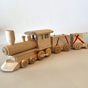May include: A wooden toy train set with a locomotive and three boxcars. The train is painted in natural wood tones with red and blue accents. The boxcars have a criss-cross pattern of red and blue lines.