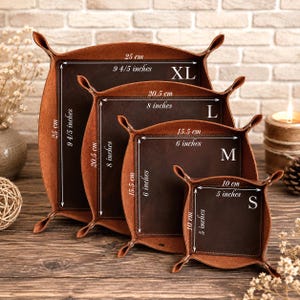 Può includere: Quattro svuotatasche quadrati in pelle marrone inseriti l'uno nell'altro, etichettati XL, L, M e S. Le dimensioni sono stampate su ciascuno: XL è 25 cm, L è 20,5 cm, M è 15,5 cm e S è 10 cm. Ogni vassoio ha angoli rialzati e base scura.