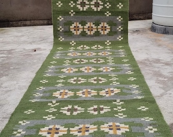 May include: A long, rectangular green rug with a repeating pattern of gray stripes and floral-like designs in yellow, white, and blue. The rug is made of a woven material and appears to be handmade.