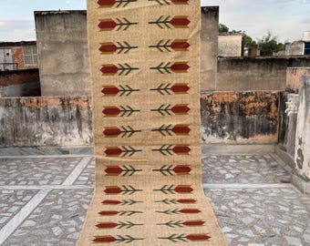 May include: Beige rectangular rug with a repeating pattern of red and green arrow-like shapes. The rug has a brown fringe at the bottom. The rug is made of a woven material.