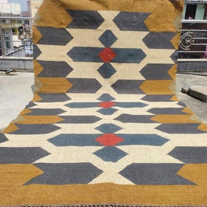 Puede incluir: Una alfombra rectangular grande con un patrón geométrico. La alfombra presenta un diseño repetitivo de formas de flecha grises, azules y doradas sobre un fondo crema. Formas de diamante rojas están intercaladas en el diseño. La alfombra está hecha de material tejido.