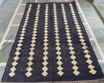 May include: A rectangular navy blue rug with a repeating pattern of diamond shapes in a lighter beige color. The rug has a fringed edge and is displayed on a gray tiled surface. The rug is a home decor item.