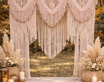 May include: A macrame wedding backdrop in a natural beige color, with intricate geometric patterns and long fringe. The backdrop is set up outdoors with pampas grass and candles, creating a bohemian aesthetic.