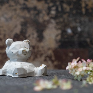 Könnte beinhalten: Eine weiße geometrische Bärenfigur mit einer abgenutzten Oberfläche sitzt auf einer grauen Oberfläche mit weißen Blumen.