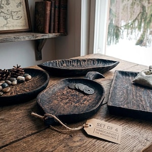 May include: A set of four dark, hand-carved wooden trays with textured surfaces. The collection includes a round bowl with pinecones and stones, an elongated oval dish, a leaf-shaped tray with coins, and a rectangular tray with a small fabric pouch.