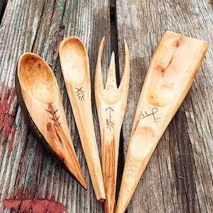 K&ouml;nnte beinhalten: Vier handgefertigte K&uuml;chenutensilien aus Holz: ein L&ouml;ffel, eine Gabel, ein Spatel und ein Servierl&ouml;ffel. Jedes St&uuml;ck ist aus hellem Holz mit dunklen Markierungen und ge&auml;tzten Designs gefertigt. Die Utensilien sind auf einer verwitterten Holzoberfl&auml;che angeordnet.