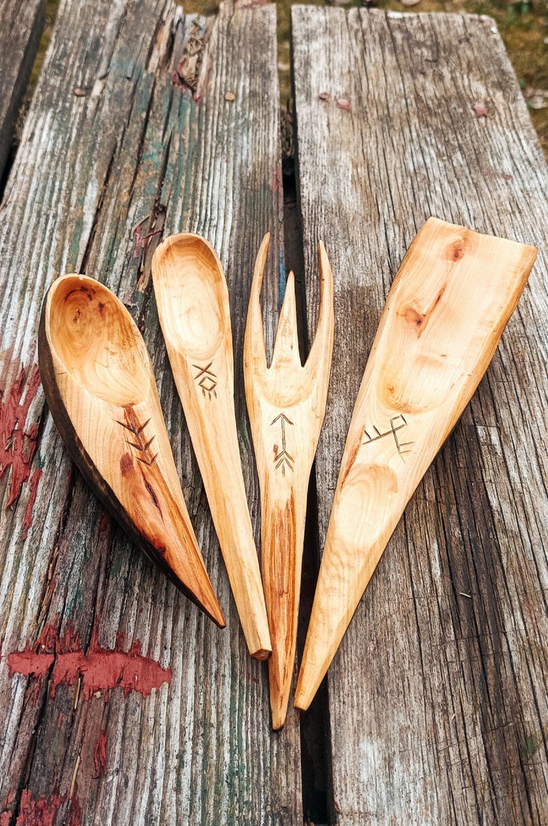 K&ouml;nnte beinhalten: Vier handgefertigte K&uuml;chenutensilien aus Holz: ein L&ouml;ffel, eine Gabel, ein Spatel und ein Servierl&ouml;ffel. Jedes St&uuml;ck ist aus hellem Holz mit dunklen Markierungen und ge&auml;tzten Designs gefertigt. Die Utensilien sind auf einer verwitterten Holzoberfl&auml;che angeordnet.