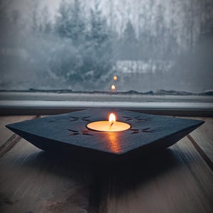 May include: A black square candle holder with a lit tea light candle in the centre. The holder has a decorative design of stars and is sitting on a wooden surface.