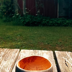 May include: A handcrafted wooden kuksa cup featuring a dark charred exterior and a natural light brown interior. The handle is carved with a leaf-like pattern and includes a rustic jute twine cord attached for hanging or carrying purposes.
