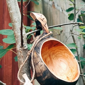 May include: A hand-carved wooden kuksa cup featuring a dark, charred exterior finish and a natural, light-colored interior wood grain. The cup includes a carved handle and a decorative wooden feather charm attached by a rustic jute twine cord.