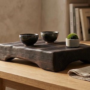 May include: A dark brown, rectangular wooden display stand with a textured, charred surface and four short feet. Two small, dark, rounded ceramic bowls and a tiny, light grey cylindrical planter containing a green moss ball sit on top.