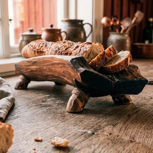 Può includere: Un vassoio da portata in legno intagliato marrone scuro con quattro gambe corte, ciascuna avvolta da spago. Il vassoio ha una consistenza naturale e ruvida e contiene una pagnotta croccante affettata con bordi carbonizzati.