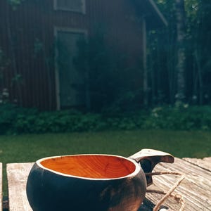 May include: A handcrafted wooden kuksa cup featuring a dark, charred exterior finish and a natural, light-colored interior wood grain. The cup includes a carved handle with an attached jute twine cord and a small, carved wooden leaf-shaped charm.