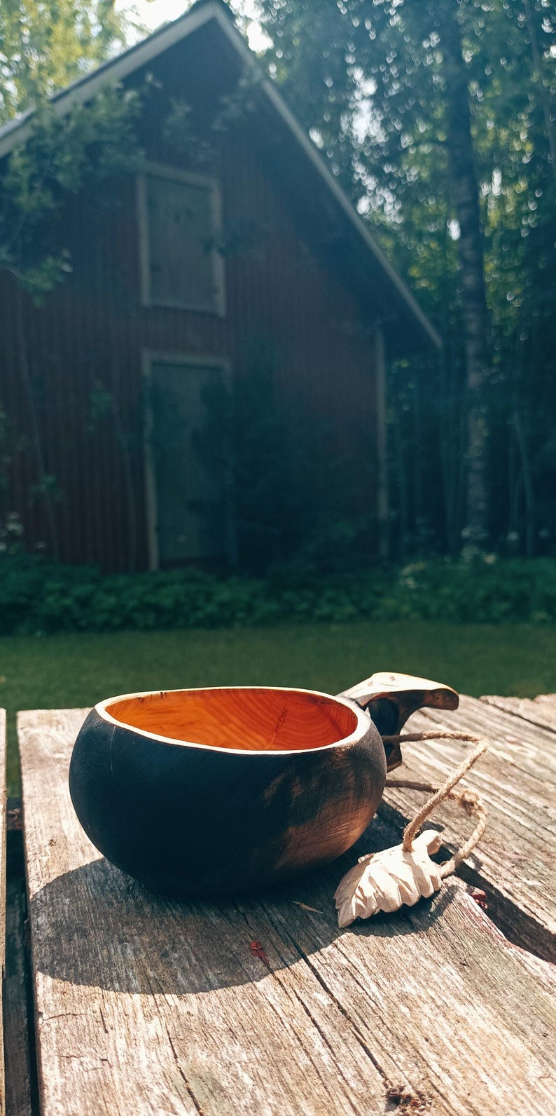 May include: A handcrafted wooden kuksa cup featuring a dark, charred exterior finish and a natural, light-colored interior wood grain. The cup includes a carved handle with an attached jute twine cord and a small, carved wooden leaf-shaped charm.