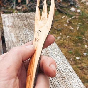 K&ouml;nnte beinhalten: Handgeschnitzte Holzgabel mit drei Zinken. Die Gabel ist hellbraun mit einem dunkelbraunen Rand und weist ein ge&auml;tztes Pfeildesign auf. Das rustikale Utensil wird vor einer verwitterten Holzoberfl&auml;che gehalten.