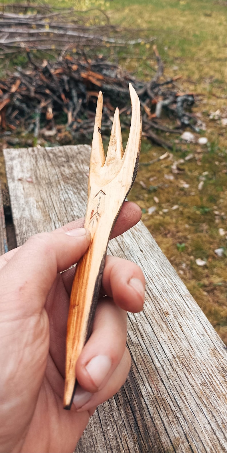 K&ouml;nnte beinhalten: Handgeschnitzte Holzgabel mit drei Zinken. Die Gabel ist hellbraun mit einem dunkelbraunen Rand und weist ein ge&auml;tztes Pfeildesign auf. Das rustikale Utensil wird vor einer verwitterten Holzoberfl&auml;che gehalten.