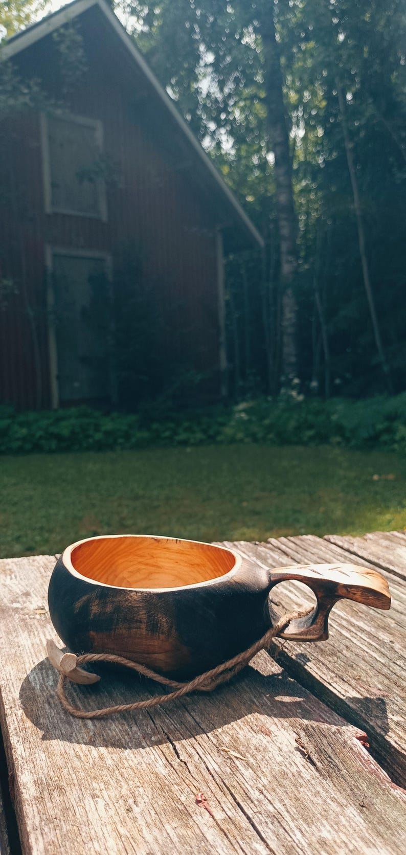 May include: A handcrafted wooden cup with a dark, charred exterior and a smooth, light-colored natural wood interior. The cup features a carved handle with a small hole and a thick, braided jute cord attached to the side for hanging or carrying.