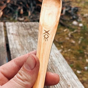 K&ouml;nnte beinhalten: Handgeschnitzter Holzl&ouml;ffel mit hellbraunem Finish. Der L&ouml;ffel hat ein geschnitztes Design in der N&auml;he des Griffs. Der unscharfe Hintergrund deutet auf ein rustikales oder nat&uuml;rliches Thema hin.