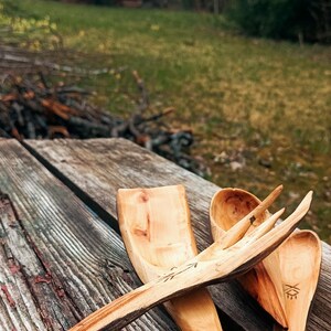 K&ouml;nnte beinhalten: Handgeschnitzte K&uuml;chenutensilien aus Holz, darunter ein L&ouml;ffel, eine Gabel und ein Spatel, liegen auf einem verwitterten Holztisch. Die nat&uuml;rliche Holzmaserung und die einzigartigen Formen der Werkzeuge werden hervorgehoben. Ein rotes Geb&auml;ude ist im Hintergrund zu sehen.