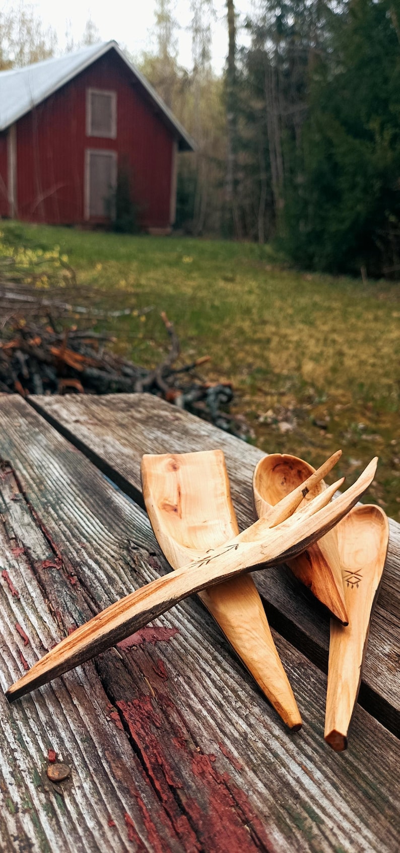 K&ouml;nnte beinhalten: Handgeschnitzte K&uuml;chenutensilien aus Holz, darunter ein L&ouml;ffel, eine Gabel und ein Spatel, liegen auf einem verwitterten Holztisch. Die nat&uuml;rliche Holzmaserung und die einzigartigen Formen der Werkzeuge werden hervorgehoben. Ein rotes Geb&auml;ude ist im Hintergrund zu sehen.