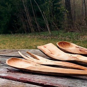 K&ouml;nnte beinhalten: Eine Sammlung handgefertigter Holzutensilien, darunter L&ouml;ffel und eine Gabel, liegt auf einer verwitterten Holzoberfl&auml;che. Die nat&uuml;rliche Holzmaserung und die warmen Farbt&ouml;ne der Utensilien kontrastieren mit dem Hintergrund im Freien.