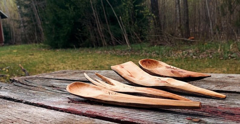 K&ouml;nnte beinhalten: Eine Sammlung handgefertigter Holzutensilien, darunter L&ouml;ffel und eine Gabel, liegt auf einer verwitterten Holzoberfl&auml;che. Die nat&uuml;rliche Holzmaserung und die warmen Farbt&ouml;ne der Utensilien kontrastieren mit dem Hintergrund im Freien.