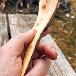 K&ouml;nnte beinhalten: Ein handgeschnitzter Holzl&ouml;ffel mit nat&uuml;rlicher, hellbrauner Oberfl&auml;che. Der L&ouml;ffel hat einen langen, schlanken Stiel und eine breite, flache Sch&uuml;ssel. Die Kanten des L&ouml;ffels sind dunkelbraun und zeigen die nat&uuml;rliche Rinde.
