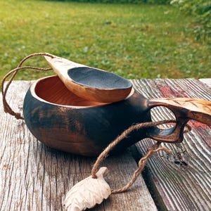 May include: A dark-stained wooden kuksa cup with a carved handle and a matching small wooden spoon resting inside. A thin twine cord is attached to the handle, featuring a small, carved wooden leaf charm dangling from the end of the string.