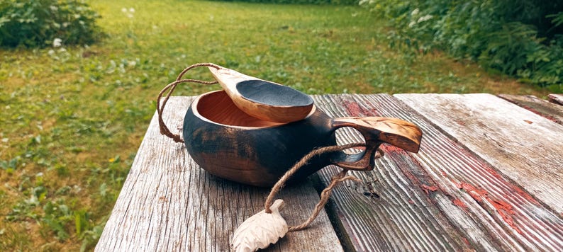May include: A dark-stained wooden kuksa cup with a carved handle and a matching small wooden spoon resting inside. A thin twine cord is attached to the handle, featuring a small, carved wooden leaf charm dangling from the end of the string.