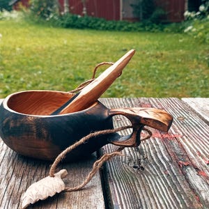 May include: A dark brown wooden kuksa cup with a smooth interior and a carved handle, featuring a matching wooden spoon resting inside. A small, carved wooden leaf charm hangs from the handle by a thin, light brown twine cord.