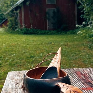 May include: A dark-stained wooden cup with a carved handle and a matching wooden spoon resting inside. The cup features a natural wood interior and a dark exterior, accompanied by a small carved leaf charm attached with a thin twine cord.