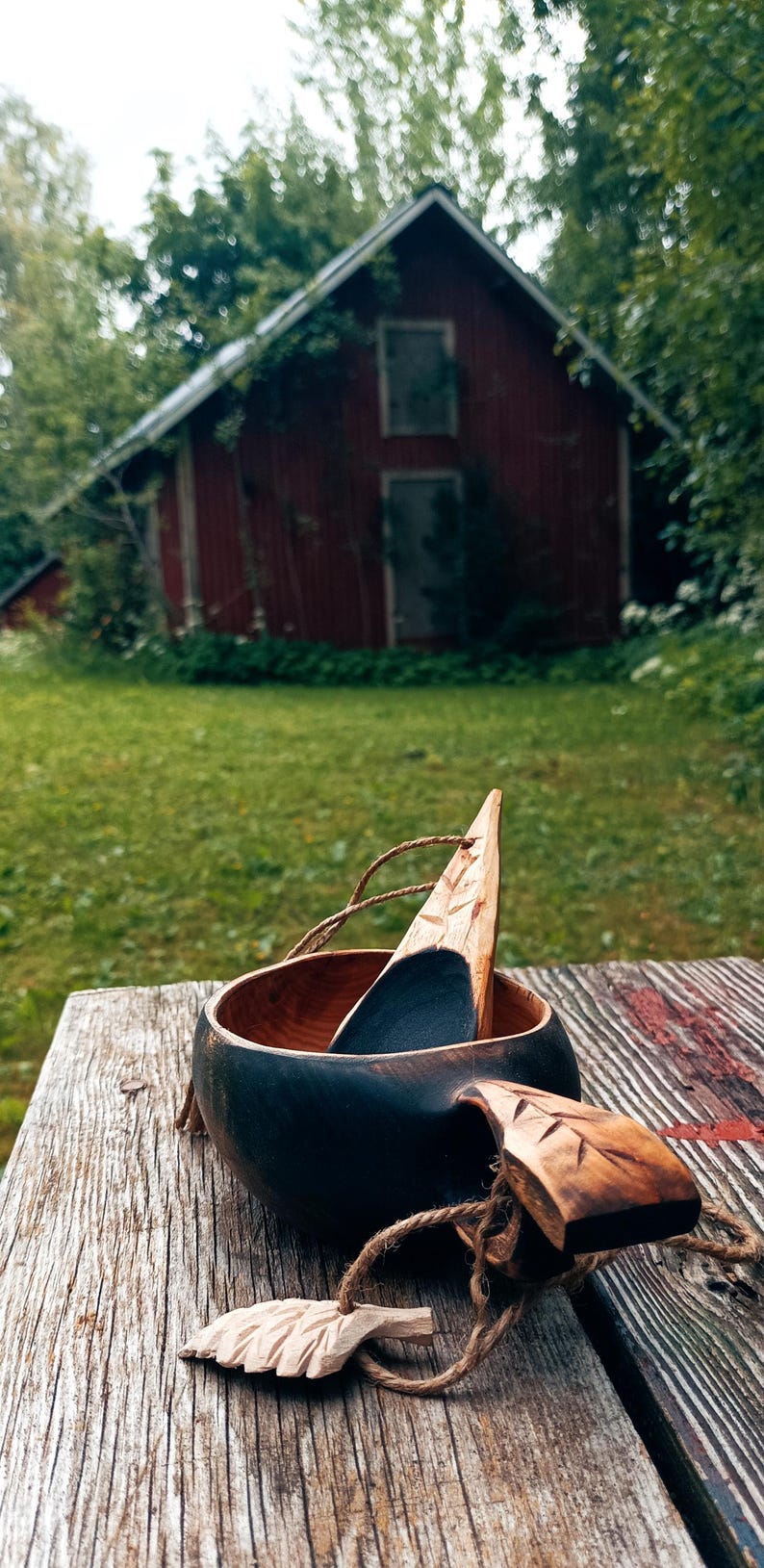 May include: A dark-stained wooden cup with a carved handle and a matching wooden spoon resting inside. The cup features a natural wood interior and a dark exterior, accompanied by a small carved leaf charm attached with a thin twine cord.