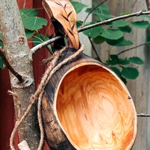 May include: A handcrafted wooden kuksa cup featuring a dark, charred exterior and a smooth, light-colored natural wood interior. The handle is carved with a leaf pattern, and a matching wooden leaf charm hangs from the cup by a rustic jute twine.