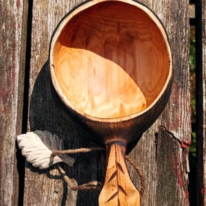 May include: A hand-carved wooden cup with a smooth, light-colored interior and a dark, charred exterior finish. The handle features a carved leaf-like pattern and is attached to a small, light-colored wooden leaf charm by a piece of brown twine.