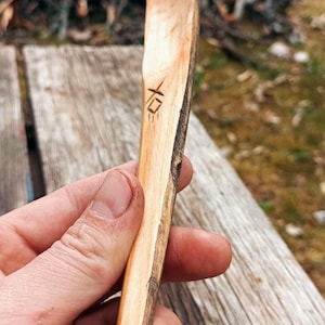 K&ouml;nnte beinhalten: Handgeschnitzter Holzl&ouml;ffel mit nat&uuml;rlicher, rustikaler Optik. Der L&ouml;ffel hat einen langen Stiel und eine abgerundete Sch&uuml;ssel mit einem strukturierten Rindenrand. Ein kleines Symbol ist in den Griff geschnitzt. Der L&ouml;ffel wird vor einem verschwommenen Hintergrund im Freien gehalten.